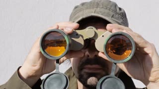 man-binoculars-sun-searching-a-man-searching-for-something-in-the-surroundings-through-a-pair-of-military-binoculars-the-orange-glass-lenses-reflect-buildings-trees-and-the-sunny-sky_vyluzcji__S0000